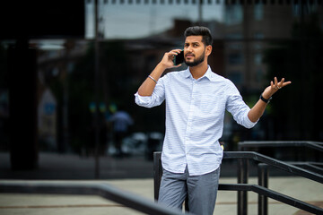 Indian handsome young businessman walking on the sunny street while holding his laptop and talking on a phone