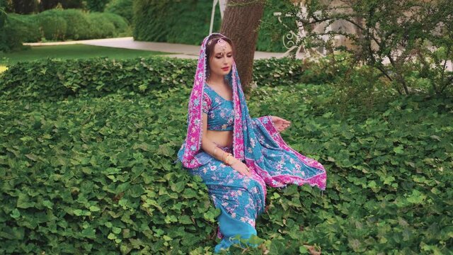 Eastern fantasy woman sits on green grass ivy, vacation meditates, enjoy nature, silence summer tropical garden park. National India blue pink dress saree, scarf on head. Silver teak tiara. Indin girl