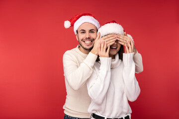 Obraz premium Photo of sweet couple in Christmas hats. Male closes her eyes up before surprise. They are in love, look blessed and smile, isolated over red background