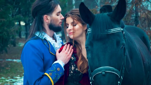 Beautiful fabulous man in love tenderly kisses happy redhaired woman. Romantic meeting of lovers in winter forest. Black horse. Blue vintage carnival camisole. Darkhaired medieval king, beard on face 