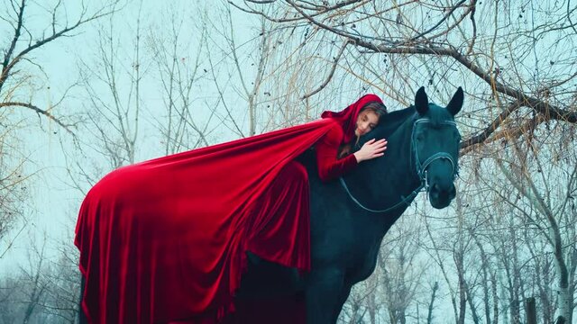 Medieval fantasy queen, horsewoman woman sittin, lies astride black horse hand strokes mare's back. Red long cloak cape. Hood on head redhaired girl princess. Bare tree branches nature winter forest.