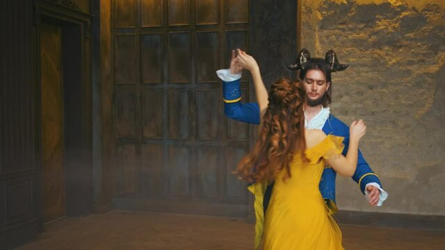 Fabulous lovers fairy tale characters beauty and beast dance romantic dance at royal ball. Family couple in costumes, blue tuxedo, yellow long vintage stylish dress. Backdrop gray medieval room 