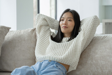 Beautiful young Asian woman, in a sweatshirt, resting on the sofa in the living room, throws her hands behind her head, closes her eyes, relaxes