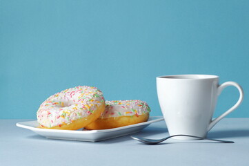 Two multi-colored donuts lie on a plate, next to it is freshly made coffee in a white cup. Composition on light blue background