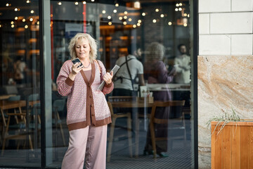 Attractive adult blonde of European appearance in a pantsuit on the streets of the city with a laptop in her hands writes a text message on a mobile phone