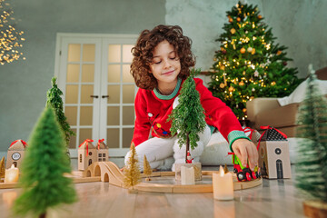 Obraz na płótnie Canvas Happy little boy with curly hair playing with wooden railroad at Christmas time
