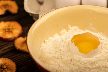 A broken white chicken egg in a bowl with wheat flour, dried apple chips and several whole eggs in a tray made of white cardboard, close-up selective focus.