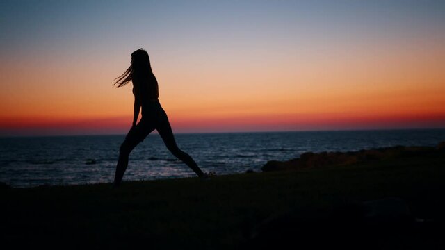 Silhouette of fitness young woman stretching legs after active workout on beach. Beautiful summer sunset over ocean. Concept of healthy and active lifestyles.