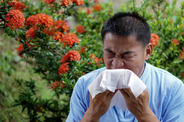 Asian man is sneezing, holding white tissue paper , open his mouth, standing beside flower trees at...