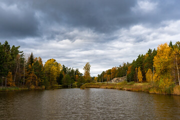 Dramatic sky with gray dark clouds on cloudy autumn day. Fall season beautiful view of small lake or pond surrounded by colorful trees. Golden yellow orange foliage. Duck house floating in the middle