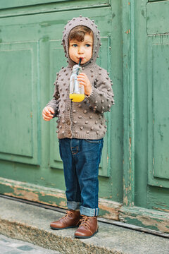 Outdoor Portrait Of Cute Toddler Girl Wearing Brown Knitted Jacket, Drinking Orange Juice From The Bottle With A Straw, Fashion For Little Children