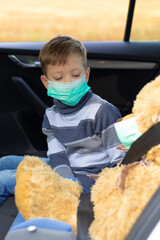 A seven year old cute boy wearing a medical mask sits in a car on a warm sunny autumn day during the coronavirus pandemic. Nearby is a toy bear. Selective focus. Portrait
