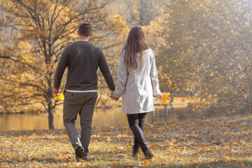 Love, leisure, nature, relationship concept. Young couple walking on meadow with yellow leaves in the park with pond and trees during autumn sunset golden ours. Bright vivid backlight filter