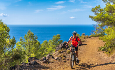 Fototapeta premium nice woman riding her electric mountain bike at the coastline of mediterranean sea on the Island of Elba in the tuscan Archipelago, in front of Porto Ferraio,Tuscany, Italy