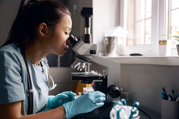 Asian lady lab assistant looks into microscope at material sample at table in hospital