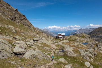 Panoramic view on Bremer mountain hut with resting tourists in sun-flooded terrace. Female hiker on Stubai high trail, Tyrol Apls.
