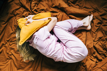 Depressed caucasian millennial woman lying alone on bed and covering head with pillow feeling afraid, top view.