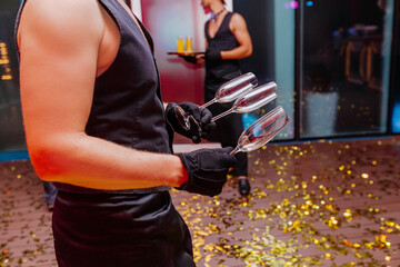 Handsome waiter holding in naked hands empty glasses for champagne. Night party celebration in club with colour disco lights. Professional restaurant serving in banquet, luxury event.