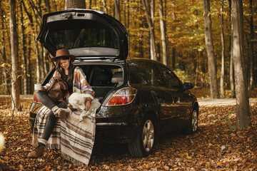 Travel and road trip with pet. Stylish happy woman traveller with cup and backpack sitting with...