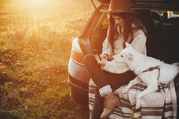 Travel and autumn road trip with pet. Stylish hipster woman in sweater holding phone and sitting...