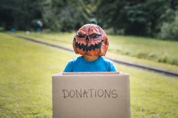 Little girl wearing a Halloween mask, holding a box asking for donations in a park.