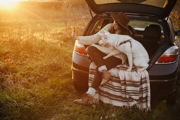 Obraz na płótnie Canvas Stylish hipster woman taking selfie photo with cute dog in car trunk in warm sunset light. Autumn road trip with pet. Young female using phone and travelling with sweet white dog. Space for text