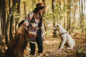 Stylish woman in hat training cute dog sitting on old stump in sunny autumn woods. Young female traveler with swiss shepherd white dog in beautiful forest. Travel and learning with pet