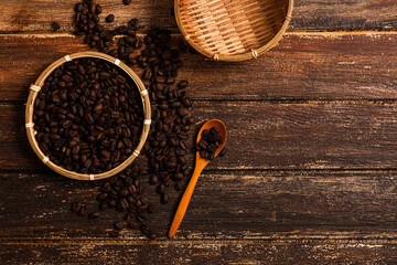 Coffee beans in a flat basket on a wood background with some coffee beans in the surrounding, and a wooden spoon and empty basket