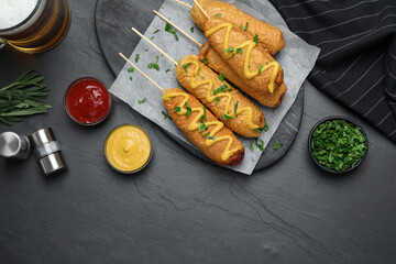 Delicious deep fried corn dogs and sauces on black table, flat lay