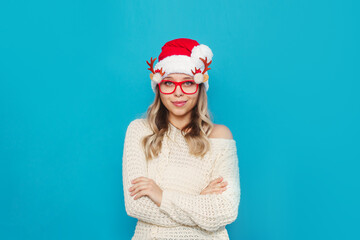 A young smiling blonde woman in glasses with deer antlers, a white knitted warm sweater and red Santa hat stands crossing her arms on her stomach isolated on color blue background. Christmas, New Year