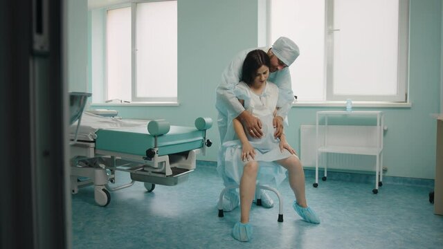 Caucasian pregnant feeling strong pain during contractions at hospital ward. Caring husband in medical clothes standing behind and massaging her back. Support and love concept.