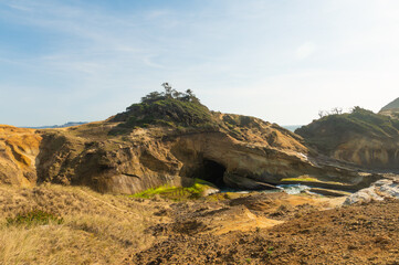 Sandy shore. There is an entrance to a small cave in the rock. Uneven hilly terrain. Blue sky with light white clouds. No people. A majestic landscape. Advertising of tourist destinations, postcard.