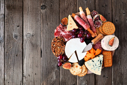 Charcuterie board of a selection of meats, cheeses and appetizers. Overhead view on a dark wood background.