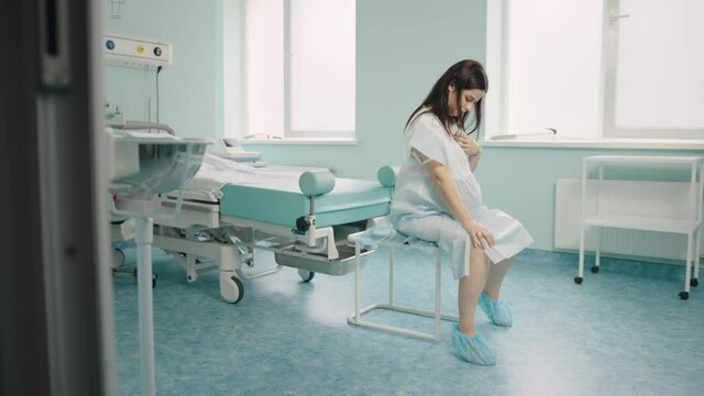 Caucasian pregnant woman with painful contraction sitting at hospital ward, touching belly and looking down. Childbirth process at hospital.