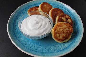 breakfast cheesecakes curd pancakes a few five pieces lie on a large blue dish plate with white sauce sour cream. Traditional dish breakfast Russian Slavic on a black background