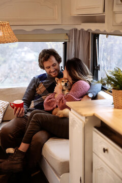 Young Couple Of Lovers Cuddling Embracing Dog Inside Camper Car. Happy Loving Man And Woman Travel In Rv Van Together With Pet. Childfree Wife And Husband On Road Trip In Motorhome Bonding Indoors