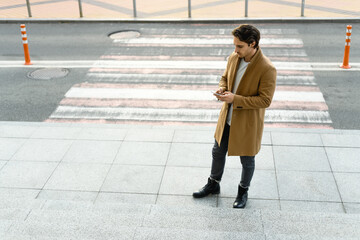 Good looking man in brown coat using smartphone near crosswalk on urban street 