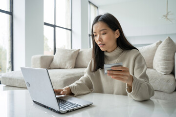 Obraz premium A young Asian woman holding a credit card and using a laptop. A female entrepreneur working from home. Online shopping, the concept of working at home