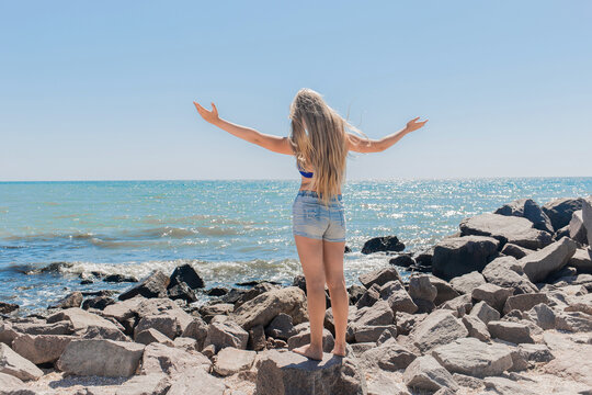A young teenage girl with long blonde hair blonde in short shorts raised and spread her hands to the sides standing on the rocks near the sea shore and the beach. The concept of freedom