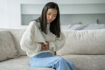 An Asian woman in a white blouse is sitting on the couch and feels a strong abdominal pain, which is called female pain or menstrual syndrome. She holds her stomach and twists in convulsions.