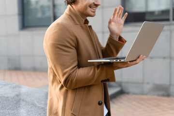 Cropped view of freelancer in coat having video call on laptop and smiling on city street 