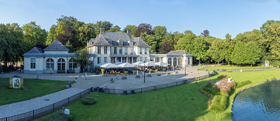 
Aerial shot of Rivierenhof castle with umbrellas in front of cafe by a pond in city park Deurne, Antwerp, Belgium. Drone aerial view