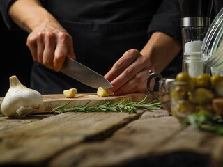 A chef in a black uniform slices garlic on a cutting board. A sprig of rosemary and olives stand on a wooden table. Preparation of salad, soup, sauce, salad, pizza, focaccia. Macro photography. - obrazy, fototapety, plakaty