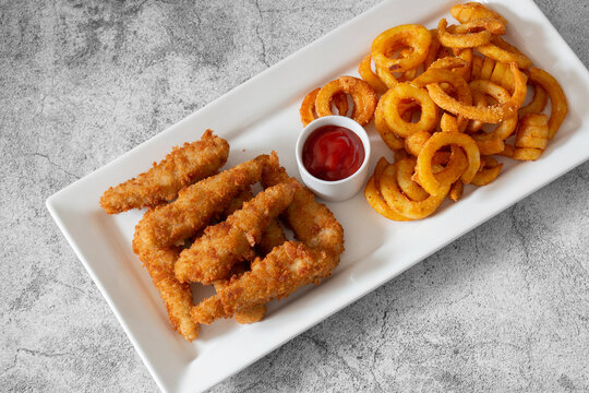 Chicken goujons, coated in breadcrumbs, with spicy curly fries and a tomato ketchup dip on a white platter.  On a concrete background