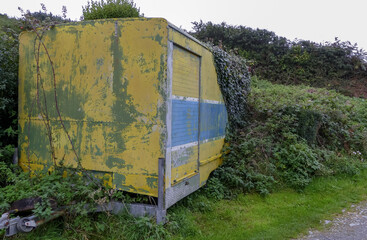 Abandoned weathered colourful industrial trailer, overgrown with ivy left by a roadside. Towing hitch Flaking yellow, blue and white paintwork. Landscape image with space for text. UK. - 463295908
