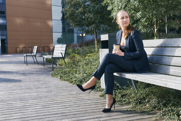 Pensive european business woman has coffee break, sits outside of office on bench with takeaway cup in hand, looks aside, wears business suit, high heels and deep neckline blouse, has ponytail.