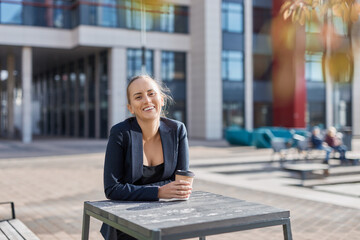 Smiling european business woman has coffee break, stands outside of office by table with takeaway coffee cup in hand, happy with project, wears ponytail, business suit and deep neckline blouse.