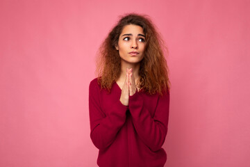 Photo of young emotional beautiful brunette curly woman with sincere emotions wearing trendy pink pullover isolated over pink background with free space and keeping hands in praying gesture and