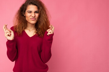 Photo shot of young attractive brunette curly woman with sincere emotions wearing trendy pink pullover isolated over pink background with empty space and keeping crossed fingers. Gesture concept