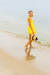 Dressing in a bright yellow sundress, holding a straw hat, looking back, a young pretty woman is walking on the beach with barefoot, relaxing..